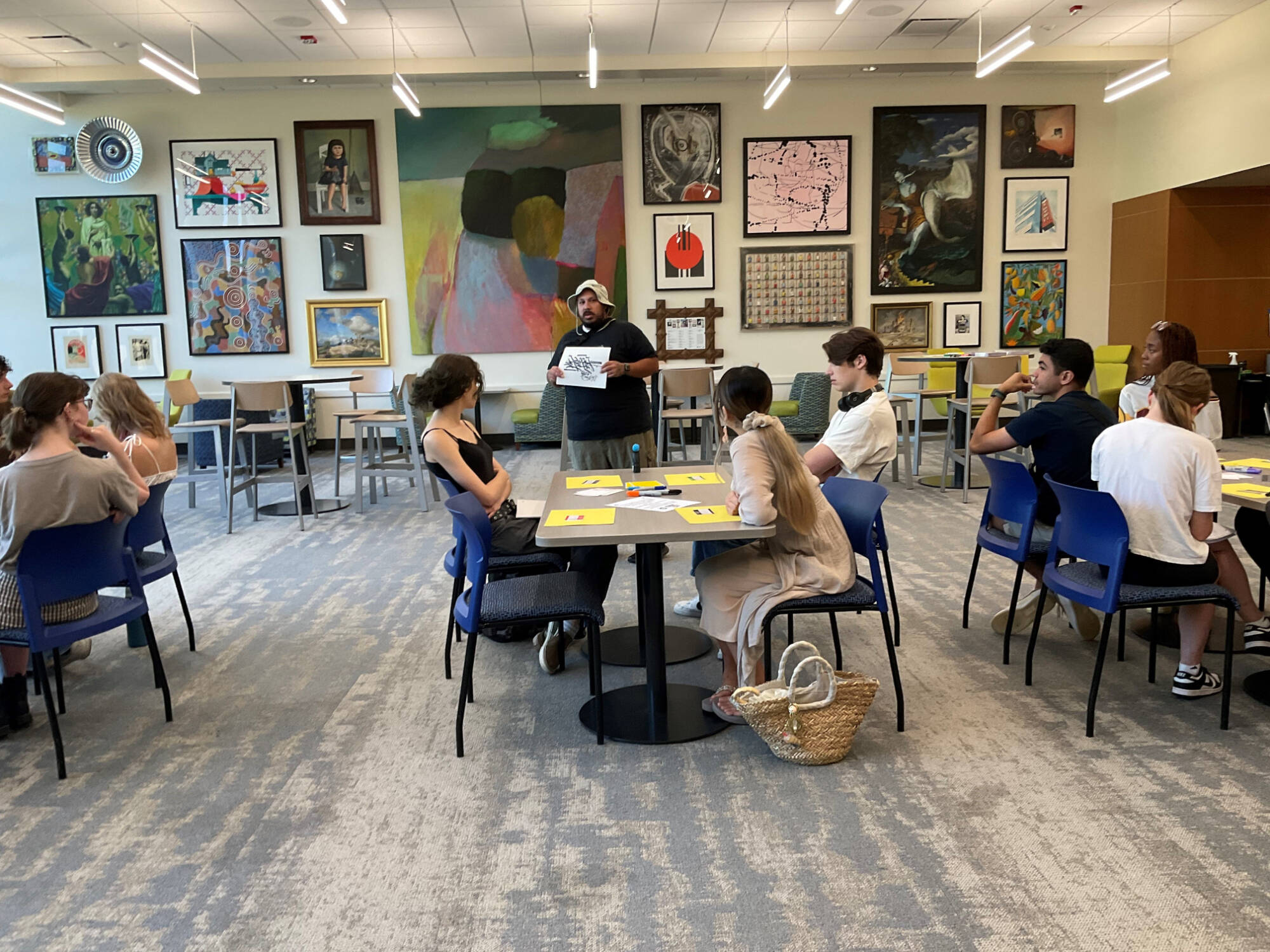 A large room with art on the wall in the background. In the midground is an artist standing and holding up an example of a mark-making project. The foreground has three tables with students sitting and listening to the artist.